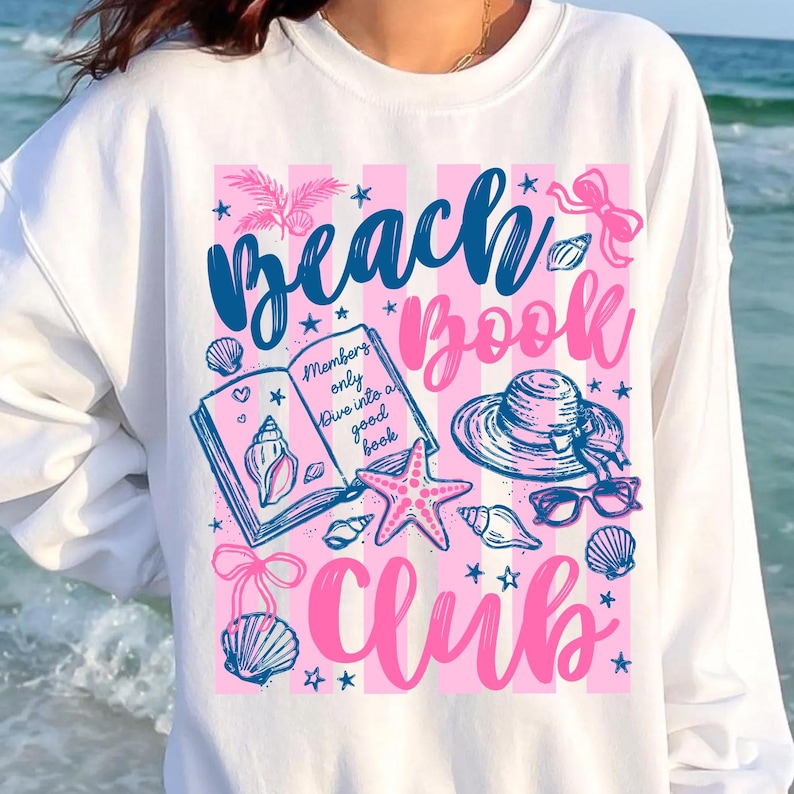 May include: White sweatshirt with a pink and blue graphic design. The design includes the words "Beach Book Club", seashells, a starfish, a hat, and sunglasses. The text "Members only dive into a good book" is also included.