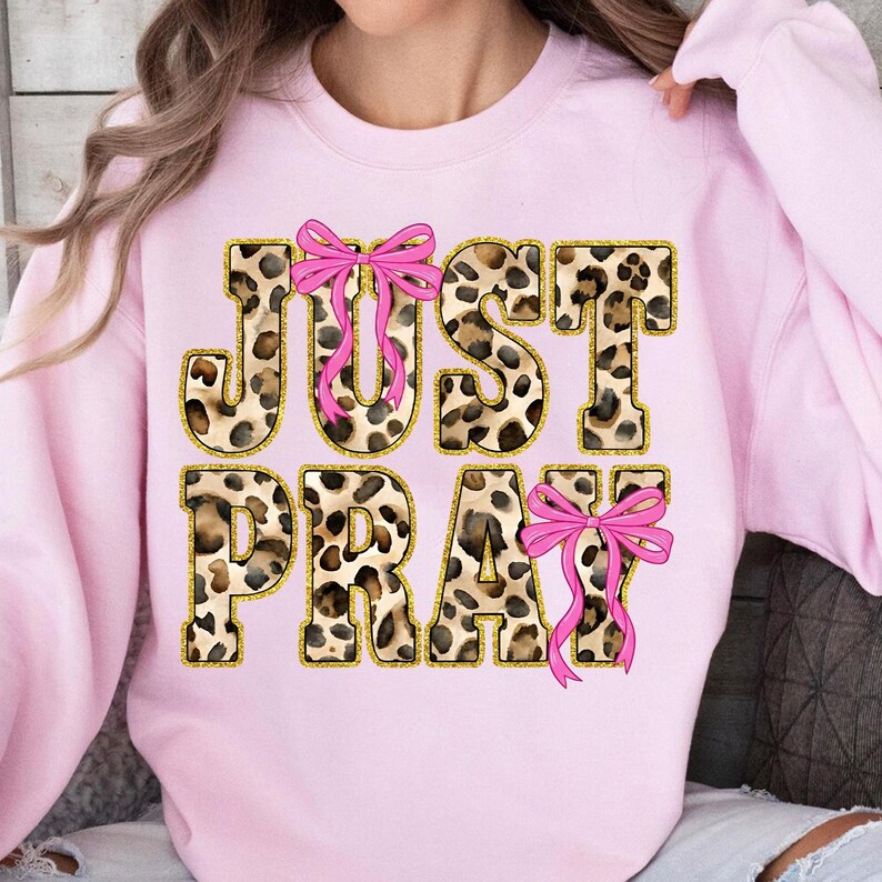 May include: A light pink sweatshirt with the words "JUST PRAY" in a leopard print design with gold trim. Pink bows are placed above the "J" and the "Y" and below the "Y".