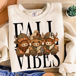 Könnte beinhalten: Ein cremefarbenes Sweatshirt mit Rundhalsausschnitt und einer Grafik von drei braunen Hochlandrindern, die gemusterte Mützen und Schals tragen. Der Text FALL VIBES ist in großen schwarzen Großbuchstaben hinter den Rindern gedruckt, die Camouflage-, Karo- und Leopardenmuster aufweisen.