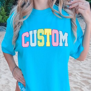May include: A turquoise t-shirt with the word "CUSTOM" in a colorful, collegiate-style font. The letters are outlined in red, with yellow and pink stripes inside. The shirt is a solid color and has short sleeves.