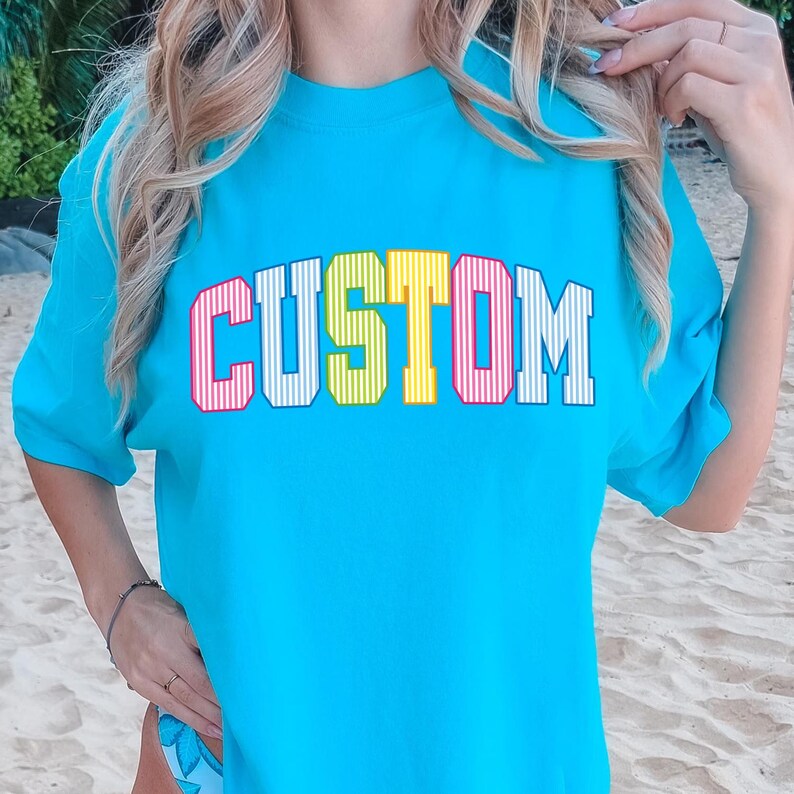 May include: A turquoise t-shirt with the word "CUSTOM" in a colorful, collegiate-style font. The letters are outlined in red, with yellow and pink stripes inside. The shirt is a solid color and has short sleeves.