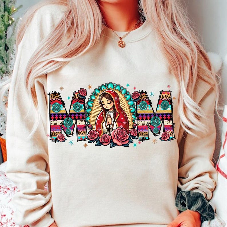 May include: Cream-colored sweatshirt with the word "MOM" in a colorful, patterned design. The letter "O" features an illustration of a woman in a red veil, surrounded by a turquoise halo and pink roses. The sweatshirt is a casual style.