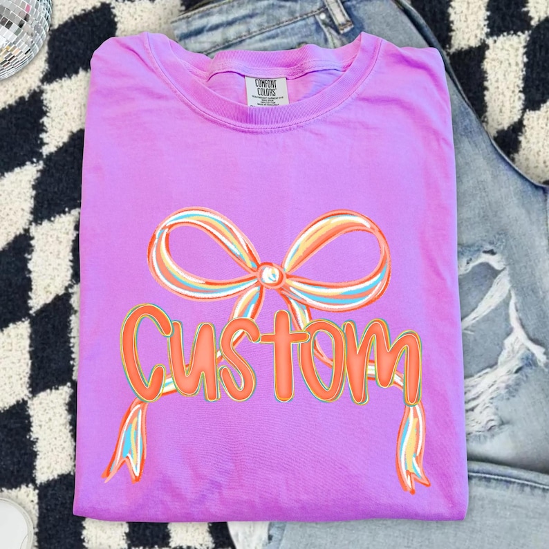 May include: A lilac t-shirt featuring a colourful bow graphic and the word "CUSTOM" in orange. The bow design incorporates shades of orange, yellow, and blue. The shirt is folded and rests on a black and white checkered surface and denim jeans.