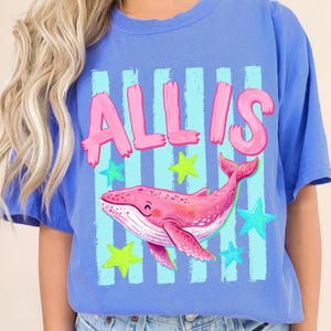 以下が含まれることがあります： ピンク色の「ALLIS」という名前がプリントされた青いTシャツ。ピンク色のクジラのイラストが描かれています。デザインには青と緑の星と縦縞が含まれています。このTシャツは柔らかい素材で作られています。