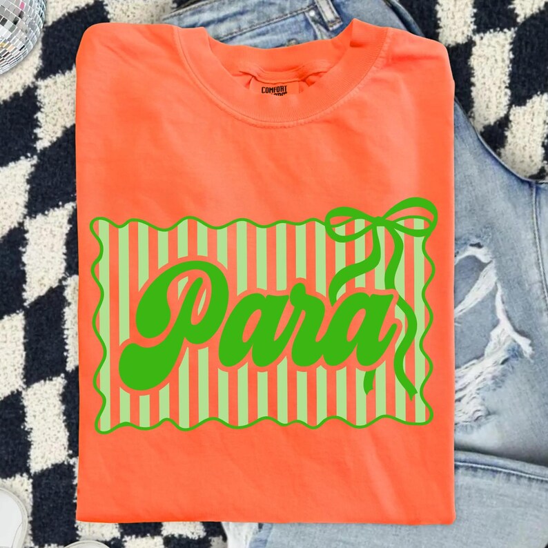 May include: Orange t-shirt with a green and light green striped design and the word "Para" in green script. The shirt is folded and displayed on a denim background.