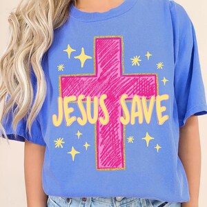 Könnte beinhalten: Ein kornblumenblaues T-Shirt mit einem großen, kritzelartigen, pinkfarbenen Kreuz, das mit goldenem Glitzer umrandet ist. Der Text "JESUS SAVE" ist in einer fetten, hellgelben Schrift mittig aufgedruckt und von kleinen gelben Sternen umgeben.