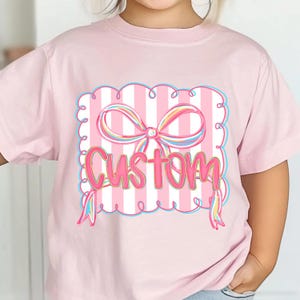 May include: A pale pink t-shirt with a graphic design. The design includes a pink and white striped background, a multi-coloured bow, and the word "Custom" in pink. The design is framed with a blue border and swirls.