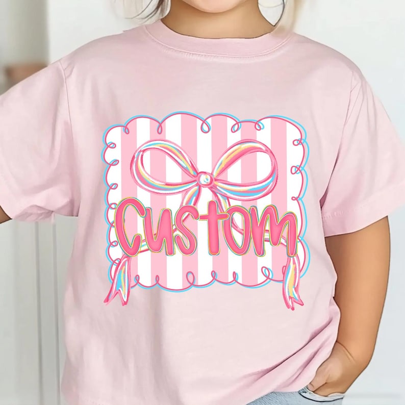 May include: A pale pink t-shirt with a graphic design. The design includes a pink and white striped background, a multi-coloured bow, and the word "Custom" in pink. The design is framed with a blue border and swirls.