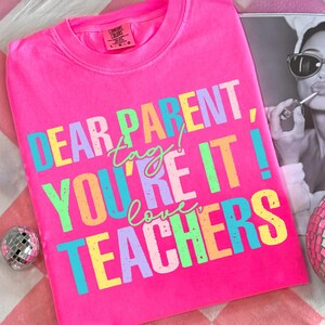 May include: Bright pink Comfort Colors t-shirt with the text "DEAR PARENT, tag! YOU'RE IT! love TEACHERS" in colorful block letters. The shirt is displayed with a black and white photo and a disco ball.