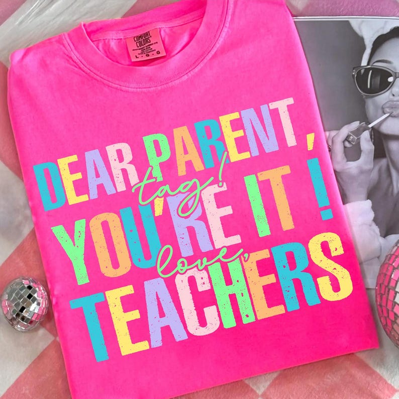 May include: Bright pink Comfort Colors t-shirt with the text "DEAR PARENT, tag! YOU'RE IT! love TEACHERS" in colorful block letters. The shirt is displayed with a black and white photo and a disco ball.