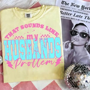 May include: A light yellow t-shirt with the phrase "THAT SOUNDS LIKE my HUSBANDS Problem" in pink and blue text. The shirt is displayed with a newspaper and a disco ball.