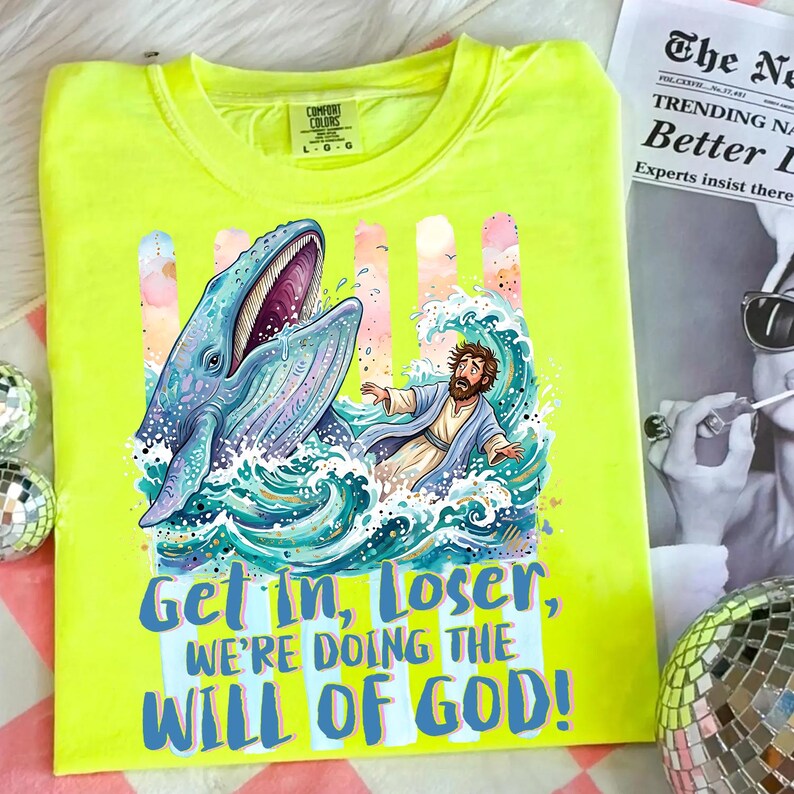 May include: A neon yellow t-shirt featuring a graphic of a whale, a man in water, and the text "Get in, Loser, We're Doing the Will of God!". The design includes blue, teal, pink, and white colors. The shirt has a crew neck and short sleeves.