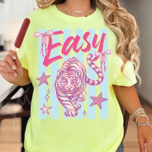 Pode incluir: Camiseta amarelo neon com um design gr&aacute;fico com a palavra "Easy" em rosa, um tigre rosa e branco e detalhes de estrelas. A camisa tem gola redonda e mangas curtas.