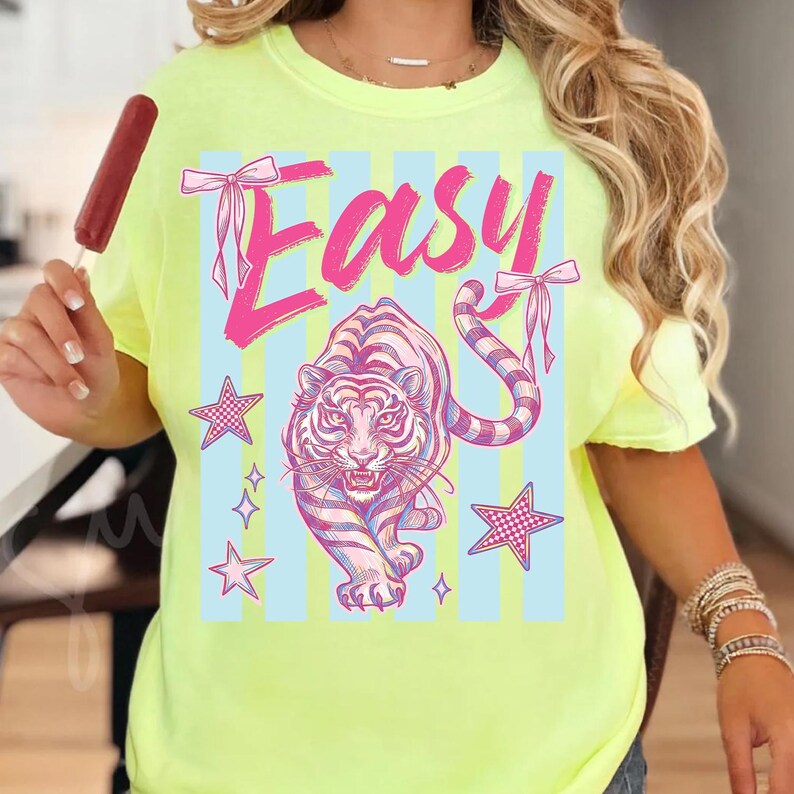 Pode incluir: Camiseta amarelo neon com um design gr&aacute;fico com a palavra "Easy" em rosa, um tigre rosa e branco e detalhes de estrelas. A camisa tem gola redonda e mangas curtas.