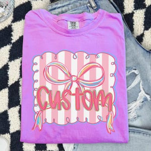 May include: A lilac t-shirt with a graphic of a pink bow and the word "CUSTOM". The design is set against a striped background with a scalloped border. The shirt is folded. The label reads "Comfort Colors".