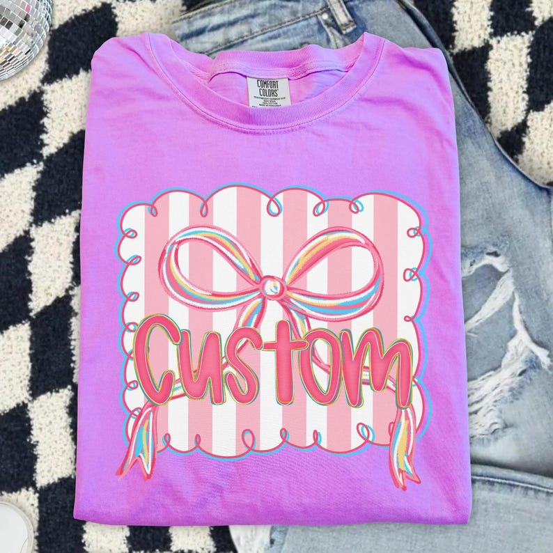 May include: A lilac t-shirt with a graphic of a pink bow and the word "CUSTOM". The design is set against a striped background with a scalloped border. The shirt is folded. The label reads "Comfort Colors".