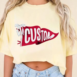 May include: A light yellow t-shirt with a red pennant design that reads "CUSTOM" in white lettering. The pennant is adorned with a white bow. The shirt is a cropped style.