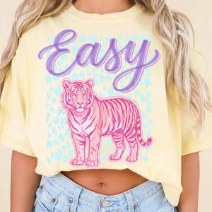 May include: Yellow t-shirt with a pink tiger illustration and the word "Easy" in purple script. The tiger is detailed with pink stripes and stands against a light blue heart-shaped pattern. The shirt is a cropped style.