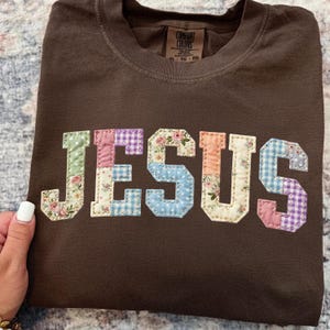 May include: A brown t-shirt with the word "JESUS" appliquéd across the front. The letters are made of various floral and patterned fabrics in pastel colors. The shirt has a crew neck and a tag that says "COMFORT COLORS".