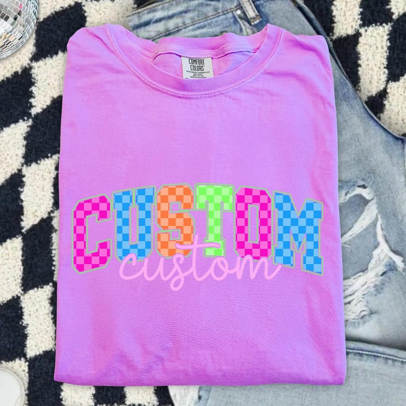 May include: A lilac Comfort Colors t-shirt with the word "CUSTOM" in a checkered pattern of pink, orange, green, blue, and a cursive "custom" underneath. The shirt is folded on a black and white checkered surface and denim jeans.