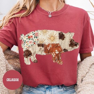 May include: Crimson-colored t-shirt featuring a patchwork pig design. The pig is composed of various fabric patterns, including floral, sunburst, and brown and white spots. The word "CRIMSON" is printed on a red circle.