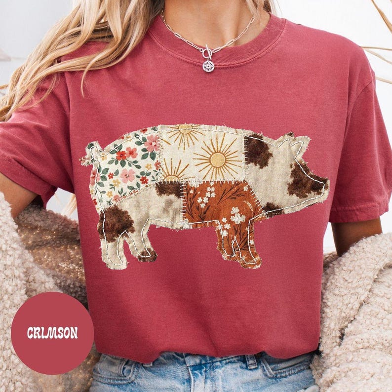 May include: Crimson-colored t-shirt featuring a patchwork pig design. The pig is composed of various fabric patterns, including floral, sunburst, and brown and white spots. The word "CRIMSON" is printed on a red circle.