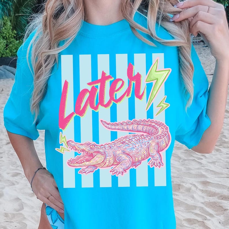 Pode incluir: Uma camiseta turquesa com a palavra "Later" em escrita rosa e um desenho de raio verde e amarelo. Abaixo do texto, h&aacute; uma ilustra&ccedil;&atilde;o detalhada de um jacar&eacute; rosa e roxo. A camiseta tem um fundo listrado.