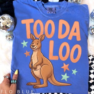 Puede incluir: Camiseta azul Flo con el texto "TOO DA LOO" en naranja. Un canguro de dibujos animados está centrado debajo del texto, con estrellas naranjas y verdes. La camiseta es de la marca Comfort Colors.