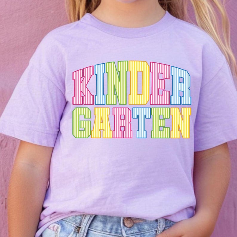 May include: A light purple short-sleeved t-shirt featuring the word KINDERGARTEN printed in a bold, block-letter font. Each letter is filled with vertical stripes in various bright colors including red, blue, green, and yellow.