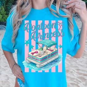May include: A turquoise t-shirt featuring a graphic of a pontoon boat on water, with the text "Pontoon Princess" in a script font. The design includes pink and white stripes and star accents.