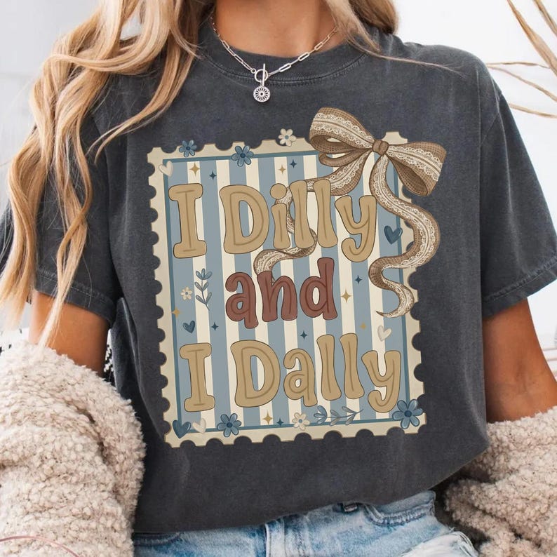 May include: Dark gray t-shirt with the text "I Dilly and I Dally" in a vintage-style design. The design features a blue and white striped background, a decorative bow, and floral accents. The shirt is a comfortable, casual wear.
