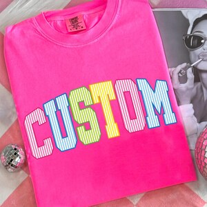 May include: A bright pink Comfort Colors crew neck t-shirt with the word "CUSTOM" in a multi-colored, striped font. The shirt has a classic fit and a tag on the upper left. A disco ball and a black and white photo are in the background.