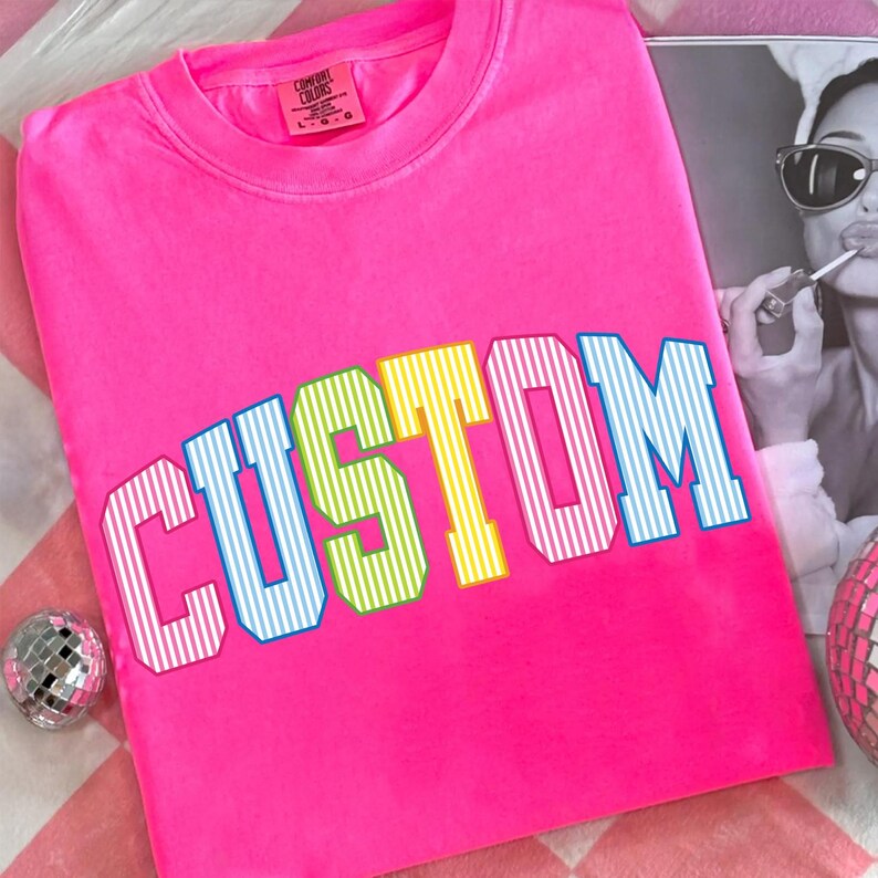 May include: A bright pink Comfort Colors crew neck t-shirt with the word "CUSTOM" in a multi-colored, striped font. The shirt has a classic fit and a tag on the upper left. A disco ball and a black and white photo are in the background.