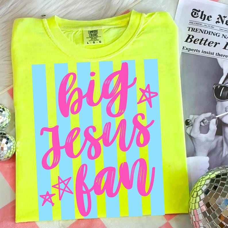 May include: A neon yellow t-shirt with the text "big Jesus fan" in pink script, set against a backdrop of light blue vertical stripes. The shirt also features star graphics. The Comfort Colors brand label is visible at the top.