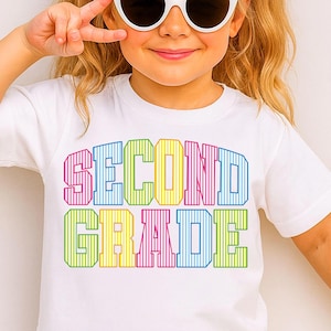 May include: A white short-sleeved t-shirt featuring the text SECOND GRADE printed in a bold, block-letter font. Each letter is filled with a vertical striped pattern in alternating colors of pink, blue, yellow, and green across two lines.
