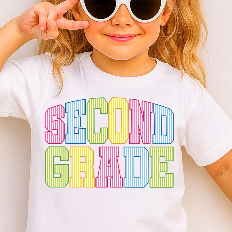 May include: A white short-sleeved t-shirt featuring the text SECOND GRADE printed in a bold, block-letter font. Each letter is filled with a vertical striped pattern in alternating colors of pink, blue, yellow, and green across two lines.