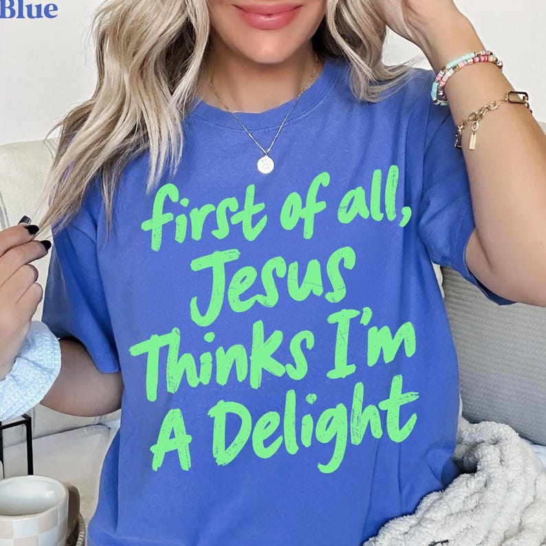 May include: A blue t-shirt with the text "first of all, Jesus Thinks I'm A Delight" in bright green. The shirt has a crew neck and short sleeves. The image shows a close-up of the shirt.