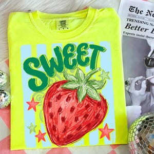 May include: A neon yellow t-shirt featuring a large red strawberry with green leaves and the word "SWEET" in green. The design includes blue vertical stripes and red and green stars.