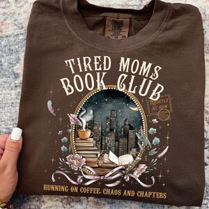 May include: A brown t-shirt with the words "Tired Moms Book Club" in white script. The graphic features a cityscape, books, coffee cup, and floral elements. The text "Running on coffee, chaos and chapters" is also included.