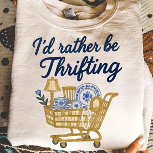 Op de afbeelding: Crèmekleurige sweatshirt met de tekst "I'd rather be Thrifting" in marineblauwe letters. Een gouden winkelwagenafbeelding is gevuld met tweedehands spullen, waaronder een lamp, servies en sieraden.