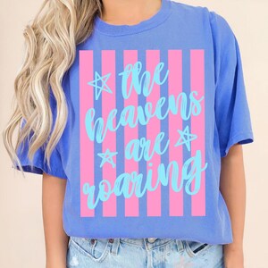 May include: Blue t-shirt with a pink and white striped design. The shirt features the text "the heavens are roaring" in a light blue script font, with star accents. The shirt is made of a soft material.