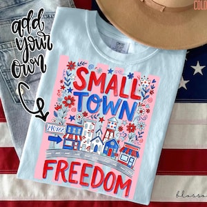Könnte beinhalten: Hellblaues Comfort Colors T-Shirt mit einem Aufdruck "SMALL TOWN FREEDOM". Das Design zeigt eine Stadtansicht mit Gebäuden, Blumen und der Postleitzahl 240988. Ein brauner Hut und eine amerikanische Flagge sind im Hintergrund.
