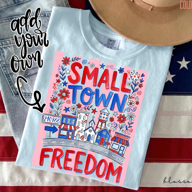 May include: Light blue Comfort Colors t-shirt with a graphic that reads "SMALL TOWN FREEDOM." The design features a town scene with buildings, flowers, and the zip code 240988. A brown hat and American flag are in the background.