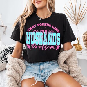 May include: Black t-shirt with the text "THAT SOUNDS LIKE my HUSBANDS Problem" in blue and pink. The shirt is worn by a person wearing blue jean shorts and a cream-colored jacket.