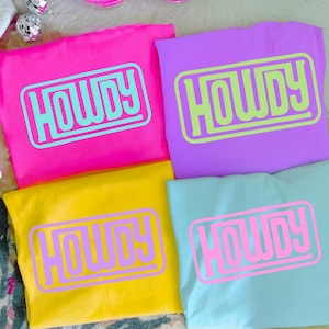 May include: Four colorful t-shirts: hot pink, purple, yellow, and light blue. Each shirt features the word "HOWDY" in a contrasting color within a rectangular frame. The shirts are neatly folded, showcasing the bold graphic design.