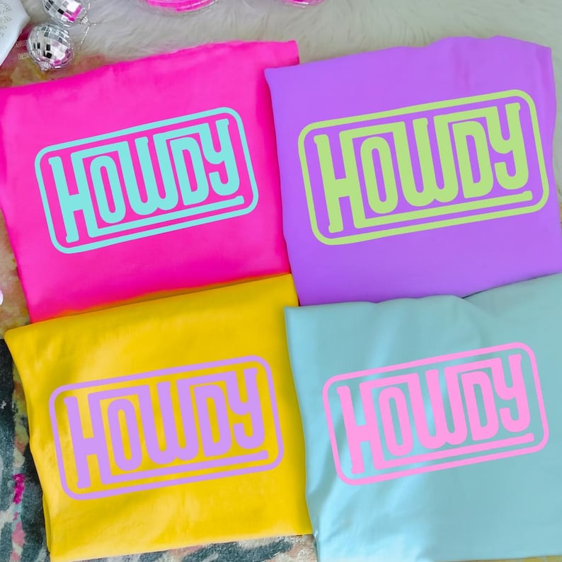 May include: Four colorful t-shirts: hot pink, purple, yellow, and light blue. Each shirt features the word "HOWDY" in a contrasting color within a rectangular frame. The shirts are neatly folded, showcasing the bold graphic design.