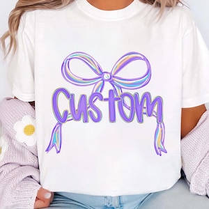 May include: White t-shirt with a purple bow design and the word "Custom" in purple script. The bow has pastel rainbow accents. This casual t-shirt is suitable for everyday wear.