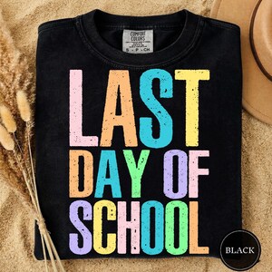 May include: Black t-shirt with the text "LAST DAY OF SCHOOL" in large, colorful, block letters. The letters are in pastel colors: pink, orange, blue, yellow, green, and purple. The shirt has a crew neck and short sleeves.