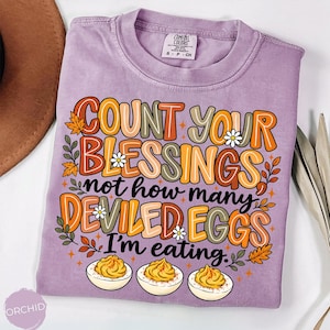 May include: A light purple short-sleeve t-shirt featuring the text "COUNT YOUR BLESSINGS not how many DEVILED EGGS I'm eating." The graphic includes illustrations of three deviled eggs, autumn leaves, and small white flowers in orange, brown, and green.