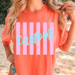 K&ouml;nnte beinhalten: Korallenfarbenes T-Shirt mit rosa Streifenmuster und dem Wort "custom" in hellblauer Schrift. Das Shirt hat kurze &Auml;rmel und einen Rundhalsausschnitt. Der Stil ist l&auml;ssig und entspannt.